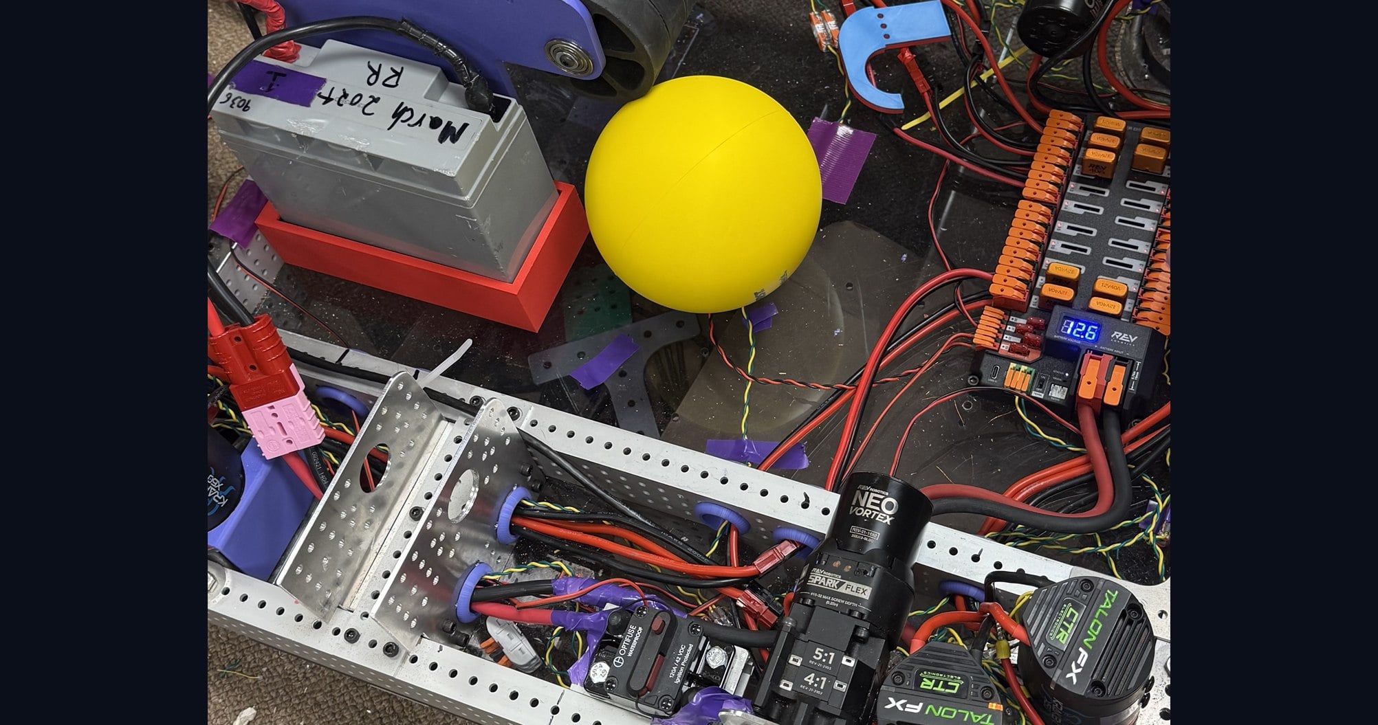 The competition robot mid-build on the shop floor: an aluminium chassis with NEO Vortex and Talon FX motors on geared drive modules, a REV power distribution hub reading 12.6 volts, a team battery, and a yellow game piece resting on the frame